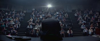 Movie still from “The Gambler” (2014), directed by Rupert Wyatt – A man sitting in front of an audience in front of a projector; Wide shot, High angle