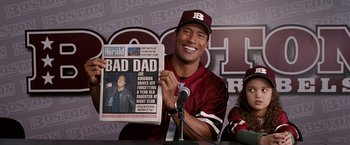 Movie still from “The Game Plan” (2007), directed by Andy Fickman – A man holding up a newspaper while sitting in front of a microphone; Medium shot, Over the shoulder angle
