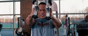 Movie still from “The Game Plan” (2007), directed by Andy Fickman – A man in a gray t - shirt is doing a workout on a machine; Close Up shot, Low angle
