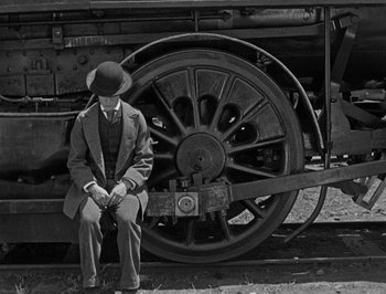 Movie still from “The General” (1926), directed by Buster Keaton – A man sitting on a bench next to a train; Wide shot, High angle