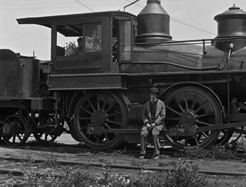 Movie still from “The General” (1926), directed by Buster Keaton – A man sitting in front of an old steam engine train; Wide shot, Low angle