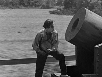 Movie still from “The General” (1926), directed by Buster Keaton – A man holding a banjo near a large object; Medium shot, Low angle