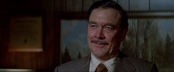 Movie still from “The Getaway” (1972), directed by Sam Peckinpah – An older man in a suit and tie smiling for the camera; Close Up shot, Over the shoulder angle