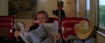 Movie still from “The Getaway” (1972), directed by Sam Peckinpah – A man sitting in a chair holding a gun and reading a newspaper; Medium shot, Over the shoulder angle