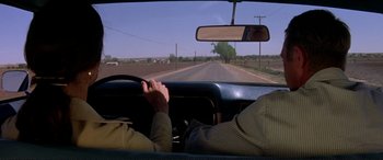Movie still from “The Getaway” (1972), directed by Sam Peckinpah – A man driving a car on a road with trees in the background; Medium shot, Over the shoulder angle