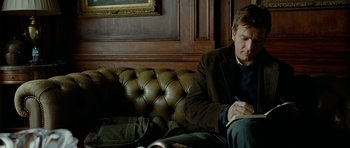 Movie still from “The Ghost Writer” (2010), directed by Roman Polanski – A man sitting on top of a couch writing on a piece of paper; Medium shot, High angle