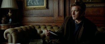 Movie still from “The Ghost Writer” (2010), directed by Roman Polanski – A man sitting on top of a couch holding a paper; Medium shot, Low angle