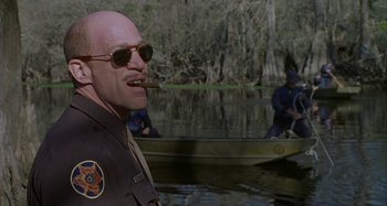 Movie still from “The Gift” (2000), directed by Sam Raimi – A man in sunglasses is smoking a cigarette in front of a boat in a body of water; Medium shot, Over the shoulder angle