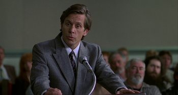 Movie still from “The Gift” (2000), directed by Sam Raimi – A man in a gray suit is speaking at a podium; Close Up shot, Over the shoulder angle