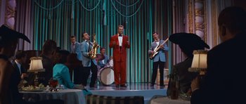 Movie still from “The Girl Can't Help It” (1956), directed by Frank Tashlin – A man in a red suit is on a stage with a group of men; Wide shot, Over the shoulder angle