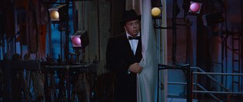 Movie still from “The Girl Can't Help It” (1956), directed by Frank Tashlin – A man in a suit and a hat standing in front of a curtain; Medium shot, Over the shoulder angle
