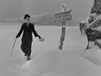 Movie still from “The Gold Rush” (1925), directed by Charles Chaplin – A man walking through the snow holding a stick; Wide shot, High angle
