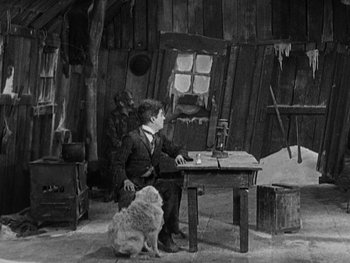 Movie still from “The Gold Rush” (1925), directed by Charles Chaplin – A man sitting at a table with a dog; Wide shot, High angle
