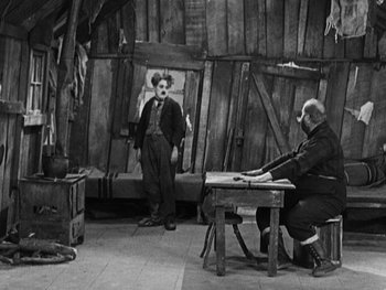 Movie still from “The Gold Rush” (1925), directed by Charles Chaplin – A man sitting at a table in a room with a clown; Wide shot, High angle