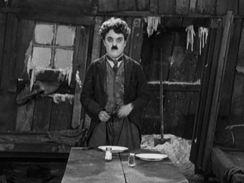 Movie still from “The Gold Rush” (1925), directed by Charles Chaplin – A black and white photo of a man with a mustache standing at a dinner table; Medium shot, Low angle