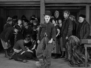 Movie still from “The Gold Rush” (1925), directed by Charles Chaplin – A group of people gathered around a man in a top hat; Wide shot, High angle