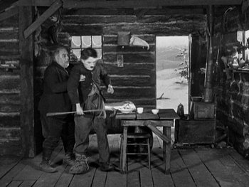 Movie still from “The Gold Rush” (1925), directed by Charles Chaplin – Two men standing next to each other in front of a table; Wide shot, High angle