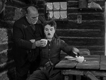 Movie still from “The Gold Rush” (1925), directed by Charles Chaplin – A man sitting at a table with another man; Medium shot, High angle