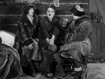 Movie still from “The Gold Rush” (1925), directed by Charles Chaplin – A man and two women sitting next to each other on a bench; Medium shot, High angle