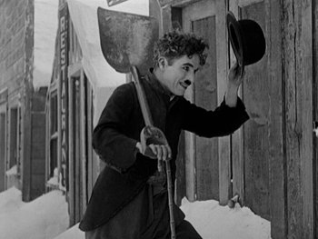 Movie still from “The Gold Rush” (1925), directed by Charles Chaplin – An old photo of a man holding a shovel and a hat; Medium shot, High angle
