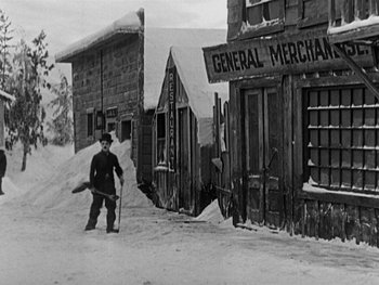 Movie still from “The Gold Rush” (1925), directed by Charles Chaplin – A man with a snow board in front of some buildings; Wide shot, Low angle