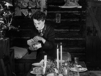 Movie still from “The Gold Rush” (1925), directed by Charles Chaplin – A man sitting at a dinner table with a plate of food in front of him; Medium shot, High angle