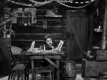 Movie still from “The Gold Rush” (1925), directed by Charles Chaplin – A man sitting at a wooden table in a room; Wide shot, High angle