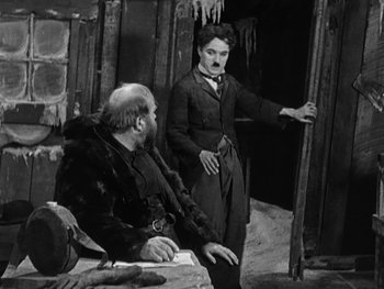 Movie still from “The Gold Rush” (1925), directed by Charles Chaplin – A man standing next to a man sitting on a bench; Medium shot, Low angle
