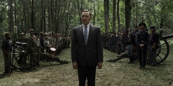 Movie still from “House of Cards” (2013), created by Beau Willimon – A man in a suit and tie standing in front of a group of people; Medium shot, Over the shoulder angle