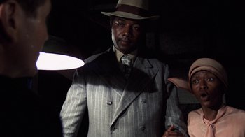 Movie still from “The Great Gatsby” (1974), directed by Jack Clayton – A man in a suit and hat standing in a dimly - lit room; Medium shot, Low angle
