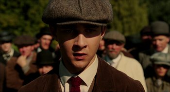 Movie still from “The Greatest Game Ever Played” (2005), directed by Bill Paxton – A young man in a hat and a tie in front of a group of people; Close Up shot, Low angle