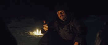 Movie still from “The Grey” (2011), directed by Joe Carnahan – A man in a black jacket and a black hat is in front of a fire; Medium shot, Low angle