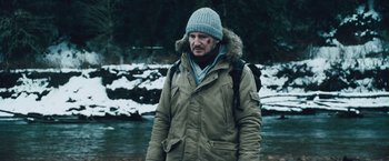 Movie still from “The Grey” (2011), directed by Joe Carnahan – A man wearing a winter coat and a hat standing in front of a river; Medium shot, Low angle