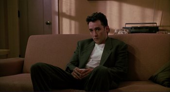 Movie still from “The Grifters” (1990), directed by Stephen Frears – A man sitting on top of a couch in a room; Medium shot, Low angle