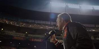 Movie still from “House of Cards” (2013), created by Beau Willimon – An older man is speaking into a microphone at a baseball game; Medium shot, Low angle