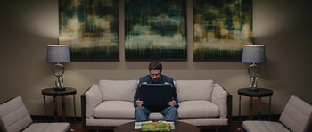 Movie still from “The Guilt Trip” (2012), directed by Anne Fletcher – A man sitting on top of a couch with a computer; Wide shot, Low angle
