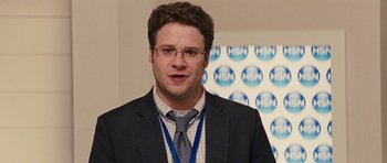 Movie still from “The Guilt Trip” (2012), directed by Anne Fletcher – A man in a suit and tie standing in front of a hsn sign; Medium shot, Over the shoulder angle