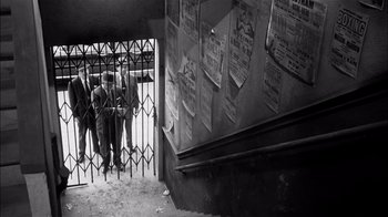 Movie still from “The Harder They Fall” (1956), directed by Mark Robson – A black and white photo of people standing in front of a building; Wide shot, High angle