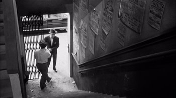 Movie still from “The Harder They Fall” (1956), directed by Mark Robson – Two men in suits and ties standing in front of a staircase; Wide shot, High angle