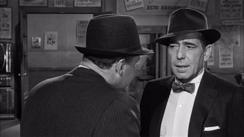 Movie still from “The Harder They Fall” (1956), directed by Mark Robson – A man in a suit and a bow tie talking to another man in a hat; Close Up shot, Over the shoulder angle