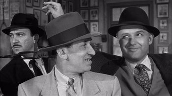 Movie still from “The Harder They Fall” (1956), directed by Mark Robson – A man in a suit and a hat is smiling; Close Up shot, Over the shoulder angle