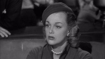 Movie still from “The Harder They Fall” (1956), directed by Mark Robson – A woman with a hat and a pearl necklace sitting down; Close Up shot, Over the shoulder angle