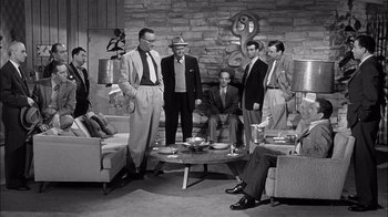 Movie still from “The Harder They Fall” (1956), directed by Mark Robson – A black and white photo of a group of men standing around a coffee table; Wide shot, High angle