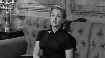 Movie still from “The Harder They Fall” (1956), directed by Mark Robson – A woman sitting on a couch in front of a wooden wall; Close Up shot, Low angle