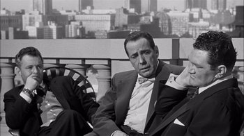 Movie still from “The Harder They Fall” (1956), directed by Mark Robson – A black and white photo of a man sitting on a bench; Medium shot, High angle