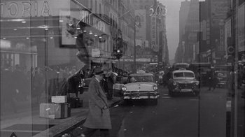 Movie still from “The Harder They Fall” (1956), directed by Mark Robson – A man in a trench coat and hat is standing on the side of the street; Wide shot, Low angle