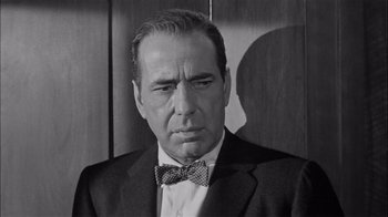 Movie still from “The Harder They Fall” (1956), directed by Mark Robson – An older man wearing a suit and a bow tie; Close Up shot, Over the shoulder angle