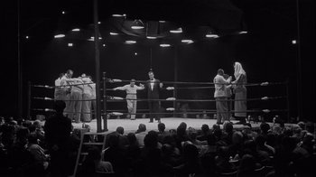 Movie still from “The Harder They Fall” (1956), directed by Mark Robson – A boxing ring filled with people in suits and robes; Wide shot, Low angle
