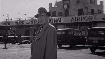 Movie still from “The Harder They Fall” (1956), directed by Mark Robson – A man wearing a hat and a coat standing in a parking lot; Medium shot, Low angle