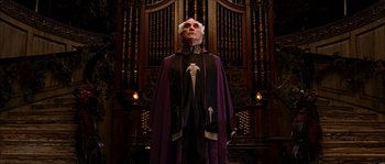 Movie still from “The Haunted Mansion” (2003), directed by Rob Minkoff – An older man in a purple robe stands in front of an organ in a church; Medium shot, Low angle